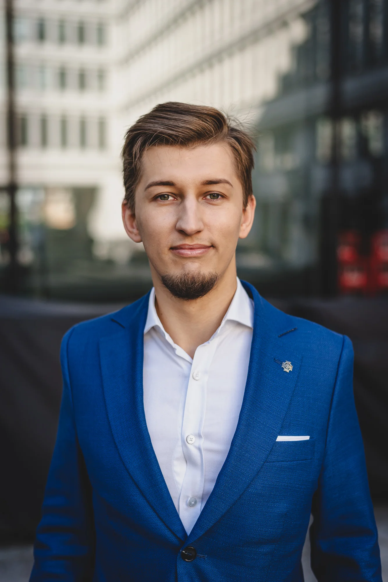 Bartosz Orzewski widoczny na zdjęciu stroi w niebieskiej marynarce na tle jasnego budynku biurowego. Lekko się uśmiecha, patrzy prosto w obiektyw