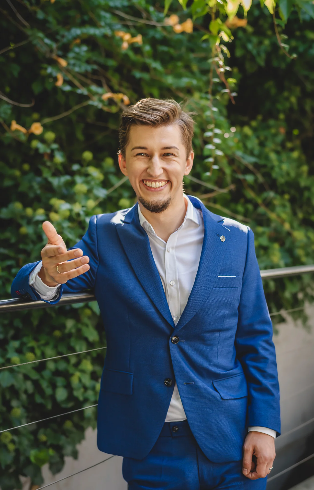 Bartosz Orzewski - młody mężczyzna ubrany w niebieską marynarkę stoi przodem oparty na metalowej barierce, na twarzy widać szeroki uśmiech, w tle widać zieleń