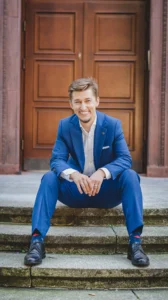 Bartosz Orzewski siedzi na schodach, w tle brązowe drzwi do warszawskiej kamienicy. Ma na sobie niebieski garnitur, bez krawata. Na twarzy widać szeroki, sympatyczny uśmiech. Łokcie umieszczone są na kolanach.