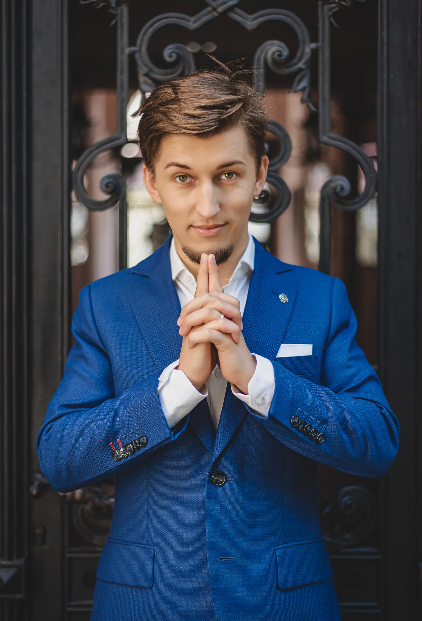 Bartosz Orzewski, ubrany w niebieski garnitur stoi na tle ozdobnej metalowej bramy w przejściu pod zabytkową kamienicą. Na twarzy widać zdecydowanie, pewność siebie. Dłonie ma złożone a palce wskazujące wyprostowane tuż pod brodą.