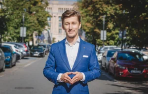 Bartosz Orzewski stroi w niebieskiej marynarce na środku warszawskieg ulicy w trakcie mówienia o niezwykłym przypadku biznesowym. W klapie ma wpięty Krzyż Harcerski, palce obydwu dłoni są złączone, na twarzy widać uśmiech