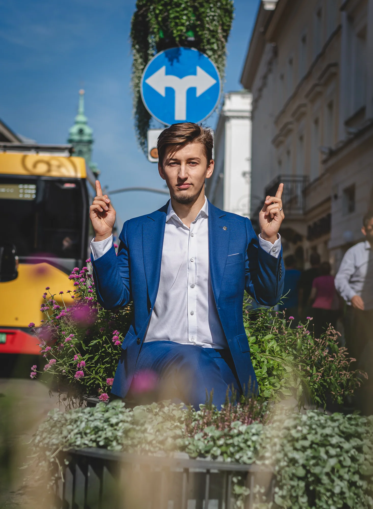 Bartosz Orzewski siedzi na donicy przy ul. Nowy Świat w Warszawie, w tle przejeżdża autobus miejski oraz przechodzą ludzie. ubrany jest w niebieską marynarkę, a palce wskazujące obydwu dłoni pokazują w górę. Nad głową widać znak nakazu jazdy w lewo albo w prawo.